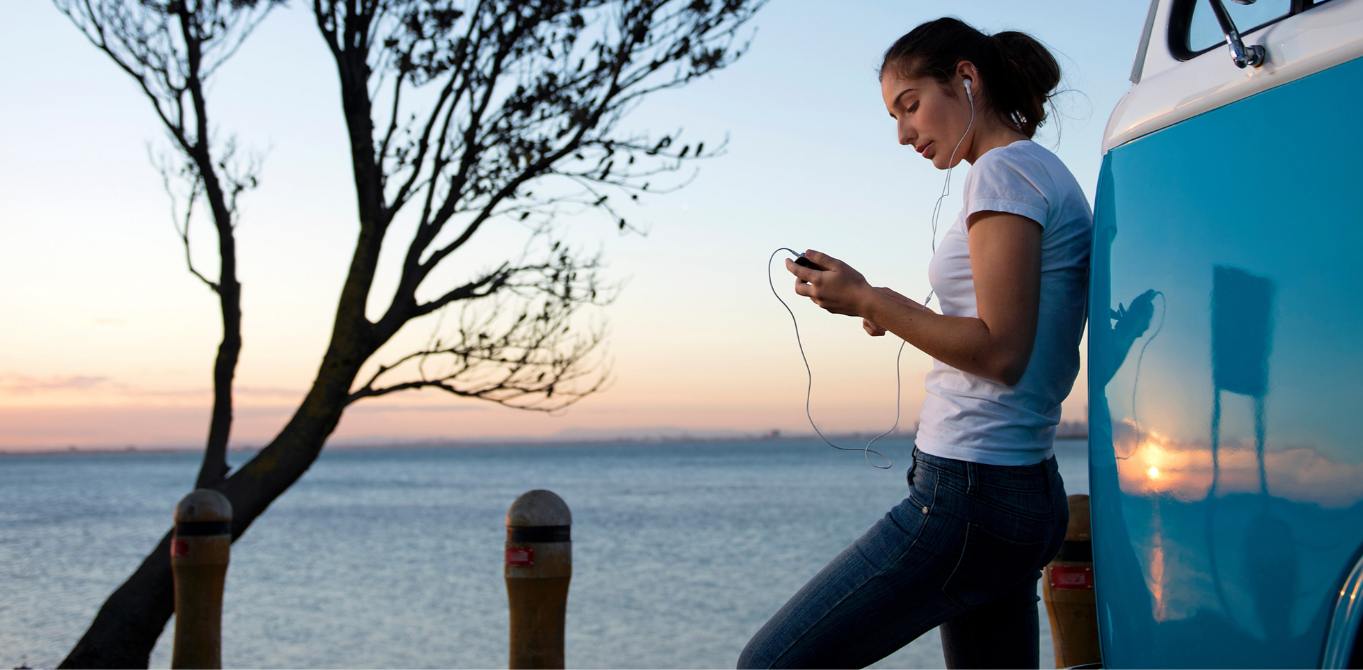A young adult woman with headphones in, leaning against a van by the water